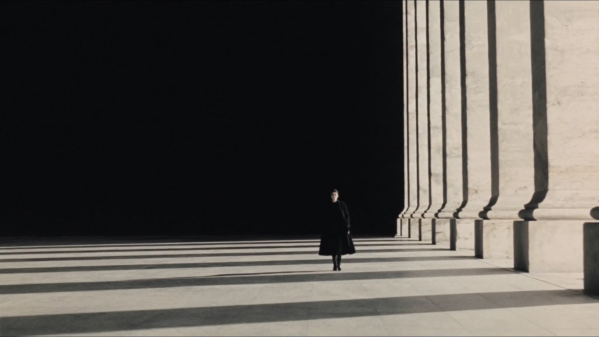 Silhouette en manteau noir marchant sous une colonnade de pierre calcaire baignée de soleil